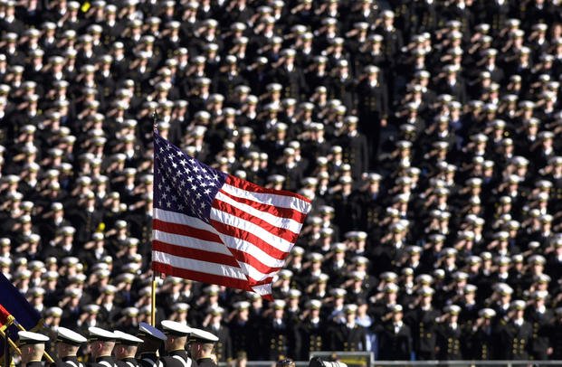 American flag flies with a background of Navy midshipmen