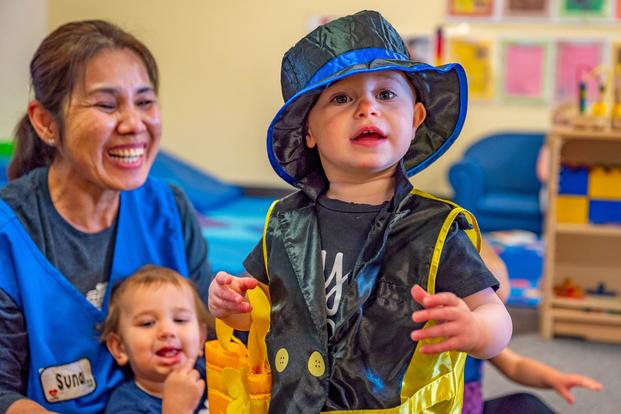 A child care worker laughs while a preschooler wears an oversized hat