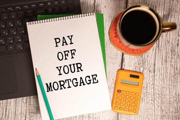 The words "pay off your mortgage" appear on a notebook resting on the wood top of a desk near office supplies and coffee.