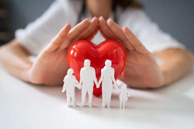 On a white tabletop, white cutouts of a family stand in the foreground in front of a heart of some kind being shielded by the hands of a person wearing a stethoscope.
