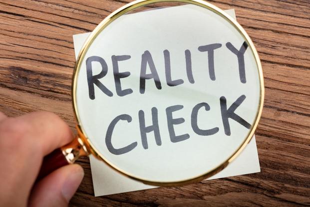 A hand holds a big, round magnifying glass over a paper with the words "reality check" printed in marker.