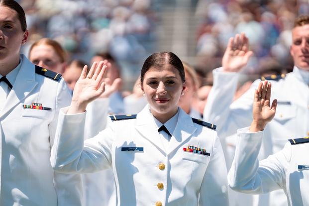 A Coast Guard Academy cadet is the central figure amid other white-uniformed cadets with their hands raised to swear the oath of office.