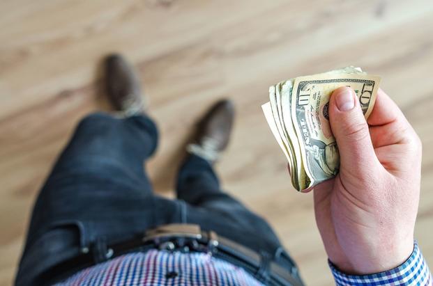 Pictured from directly above, a hand holds folded cash in the foreground and a belt, jeans and a wood floor appear blurry.