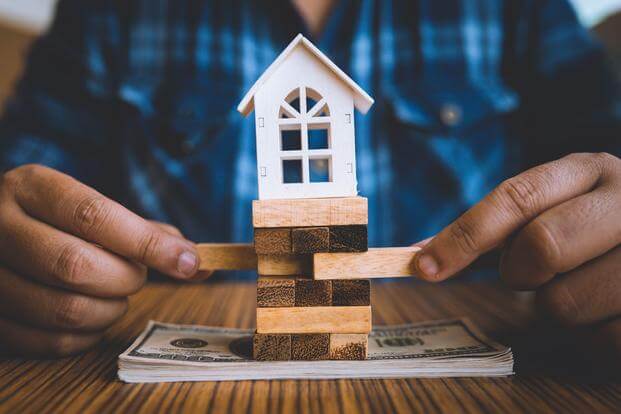 Hands pull out pieces from a Jenga wood puzzle set on a stack of cash with a toy house balancing on top.