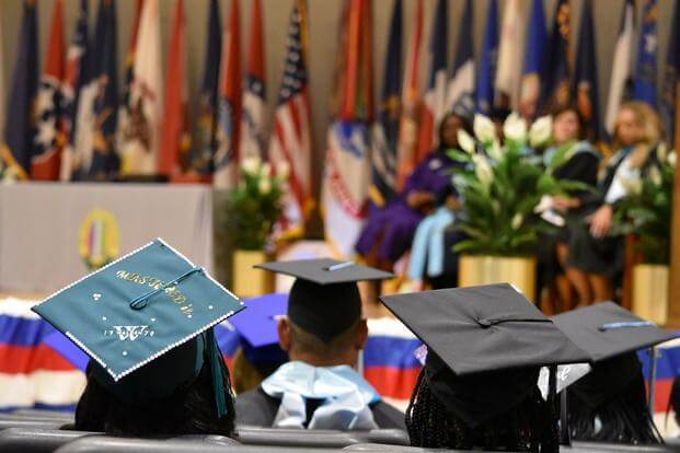 Graduate mortarboards, one decorated, pictured from the rear with a row of flags representing the military services on a stage in the foreground.