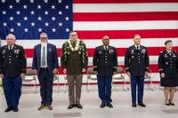 Six men and women, most in uniform, stand in a row in front of an American flag draped as a backdrop.