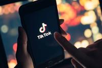 In a darkened room, a pair of hands hold and prepare to tap a phone screen displaying the TikTok logo.