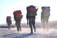 Special Forces candidates walk on a dusty road with backpacks and guns.