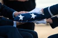 A uniformed airman wearing white gloves hands a folded flag to a seated person.