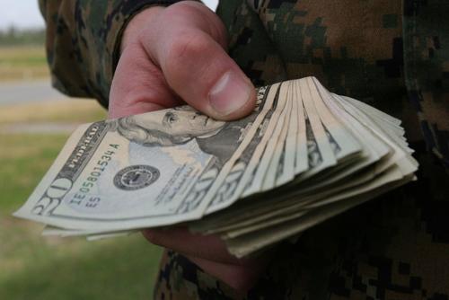 A hand of a person wearing a Marine Corps digital camouflage uniform fans out a stack of $20 bills.