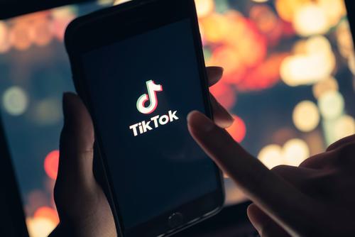 In a darkened room, a pair of hands hold and prepare to tap a phone screen displaying the TikTok logo.